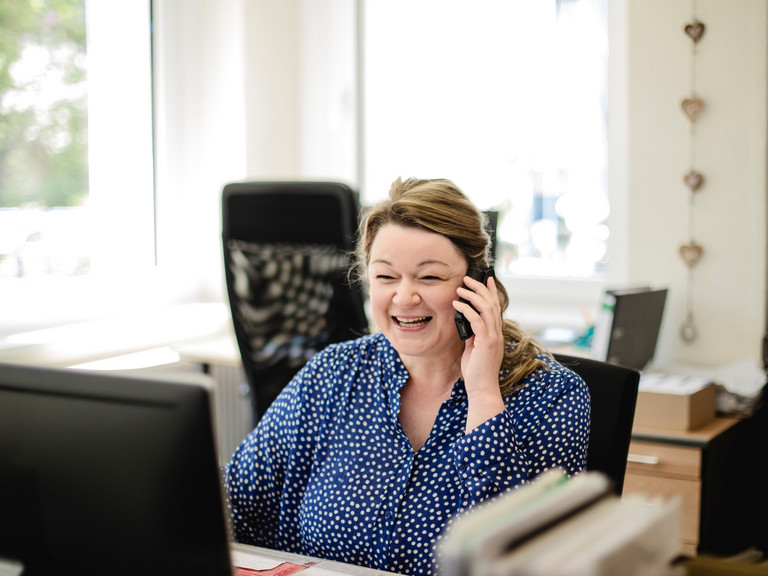 Eine Verwaltungsmitarbeiterin sitzt an einem Schreibtisch und blickt mit einem Telefonhörer in der Hand auf einen Monitor in der Verwaltung der Senioreneinrichtung Immanuel Haus Ecktannen in Waren (Müritz)