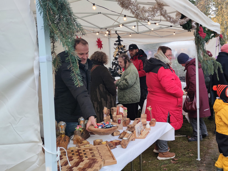 Mehrere Personen in bunten jacken stehen an einem Stand mit Holzarbeiten auf dem Weihnachtsmarkt des Immanuel Haus Ecktannen Mehrere Personen in bunten jacken stehen an einem Stand mit Holzarbeiten auf dem Weihnachtsmarkt des Immanuel Haus Ecktannen